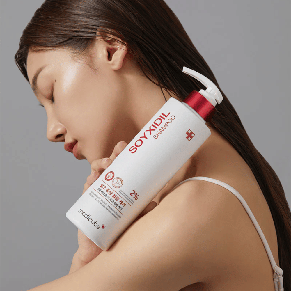 Kvinde med langt hår læner hovedet mod en flaske Medicube Soyxidil Shampoo, som styrker hovedbunden og modvirker hårtab.