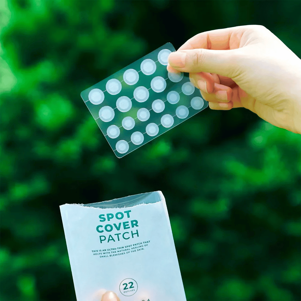 En hånd holder et ark med Spot Cover Patches, som er gennemsigtige og afrundede. I baggrunden ses et sløret grønt miljø. Emballagen viser en del af emballagen med teksten "SPOT COVER PATCH" og information om, at produktet indeholder 22 plastre, der hjælper med at hele små hudområder.