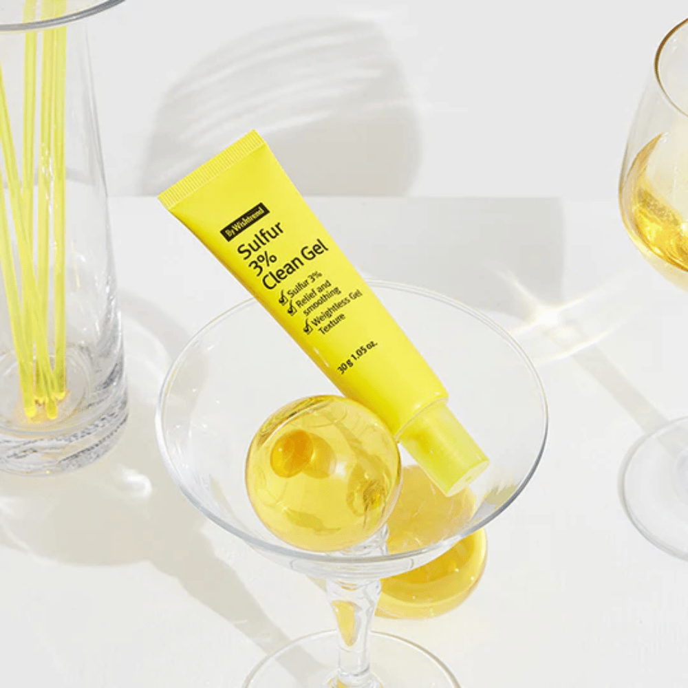 By Wishtrend Sulfur 3% Clean Gel i en gul tube placeret i et cocktailglas med en gul sfære, omgivet af elegante glas på en hvid baggrund.