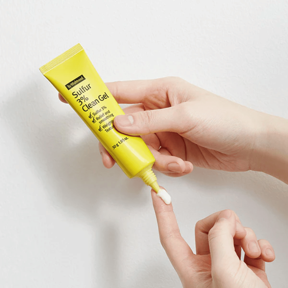 Nærbillede af hænder, der påfører By Wishtrend Sulfur 3% Clean Gel, en gul tube med hvid gel, mod en hvid baggrund.