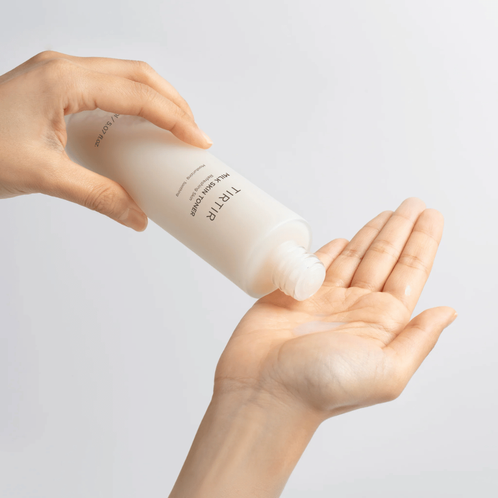Billedet viser en person, der hælder en mælkeagtig væske fra en flaske mærket "TIRTIR Milk Skin" på håndfladen. Produktet ser ud til at være en hudtoner eller fugtgivende lotion.
