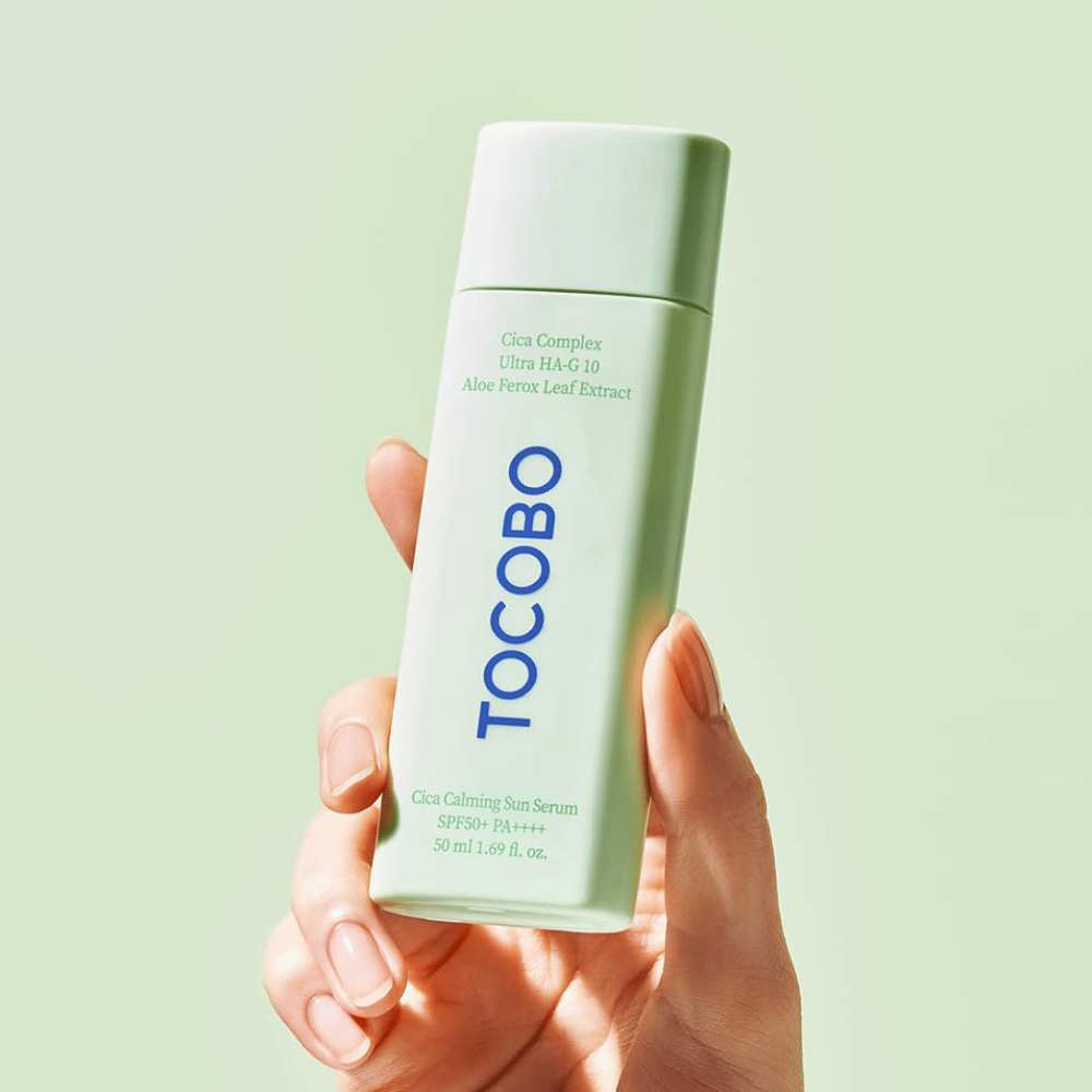 Hånd holder i TOCOBO Cica Calming Sun Serum med mintgrøn emballage og blå tekst mod lysegrøn baggrund.