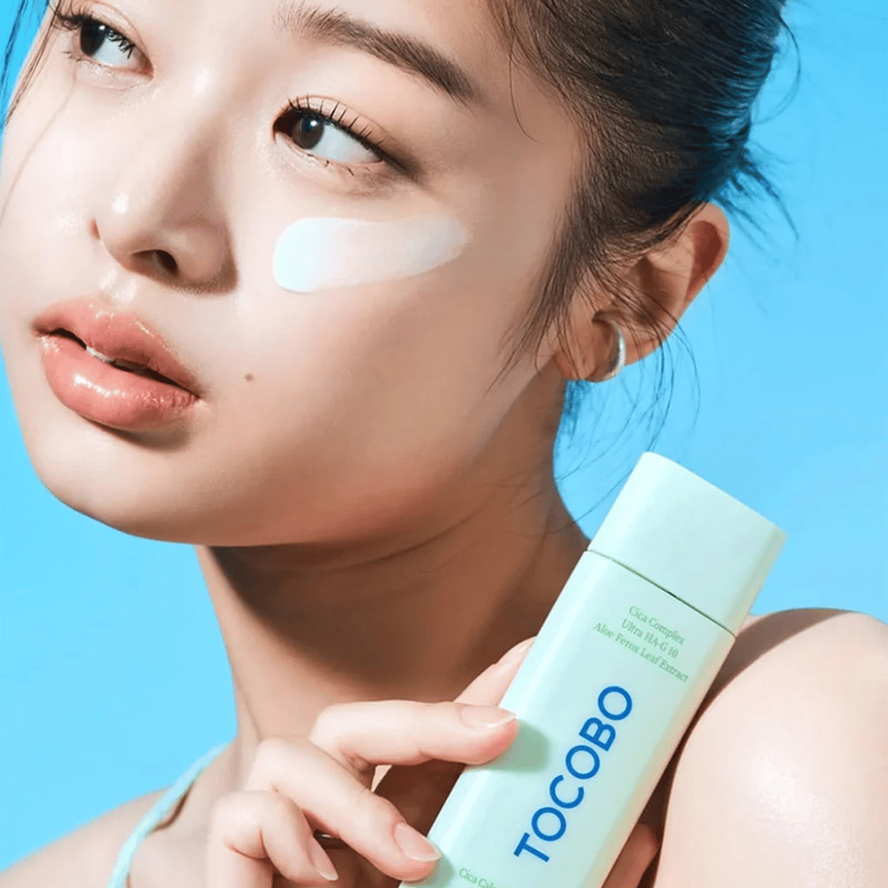 Model med solcreme under øjet holder i TOCOBO Cica Calming Sun Serum i mintgrøn emballage mod blå baggrund.