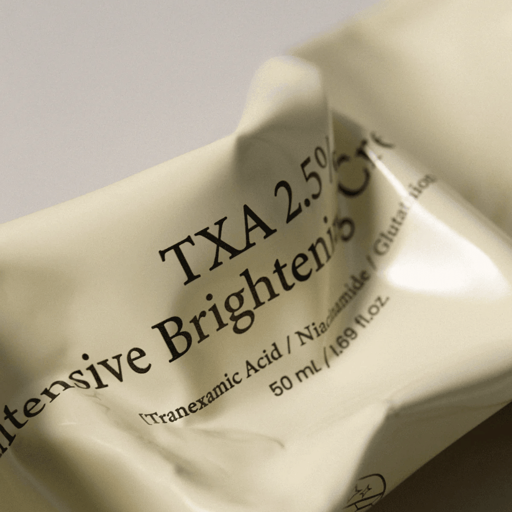 Nærbillede af AXIS - Y TXA 2.5% Intensive Brightening Cream hvor det bløde og minimalistiske tubedesign er i fokus. Billedet fremhæver produktnavn, Tranexamic Acid og Niacinamide samt den elegante beige emballage.