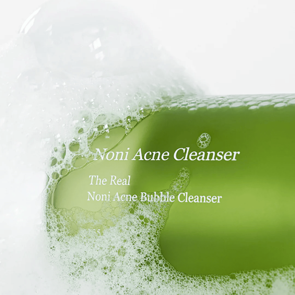 Flasken med The Real Noni Acne Bubble Cleanser er i fokus og omgivet af rigeligt med skum. Den friske grønne farve signalerer naturlige ingredienser, perfekt til at rengøre og bekæmpe akne. Et ideelt produkt til en ren og klar hud.