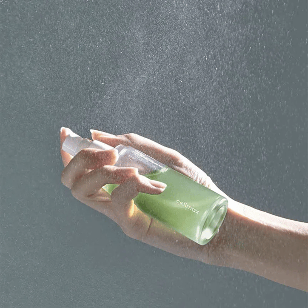 En hånd holder Celimax Noni Mist sprayflaske og påfører produktet i en fin, jævn spray på huden. Flasken er grøn og produktet markedsføres som "The Real Noni Energy Ampule Mist" for at give energi og fugt til huden.