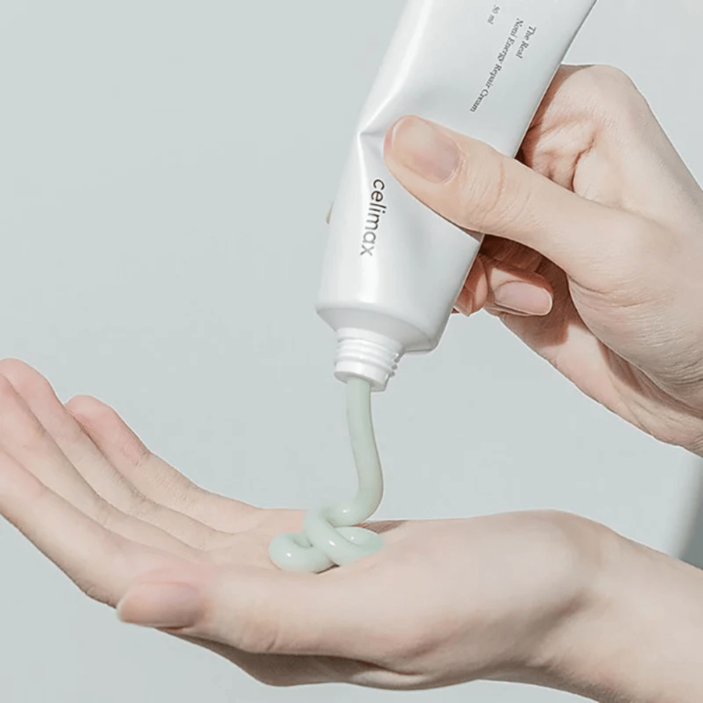 En hånd holder en hvid tube med Celimax Noni Energy Repair Cream, og cremen presses ud i en jævn stråle på håndfladen. Cremen er lysegrøn og markedsføres til at give næring og genoprette huden.