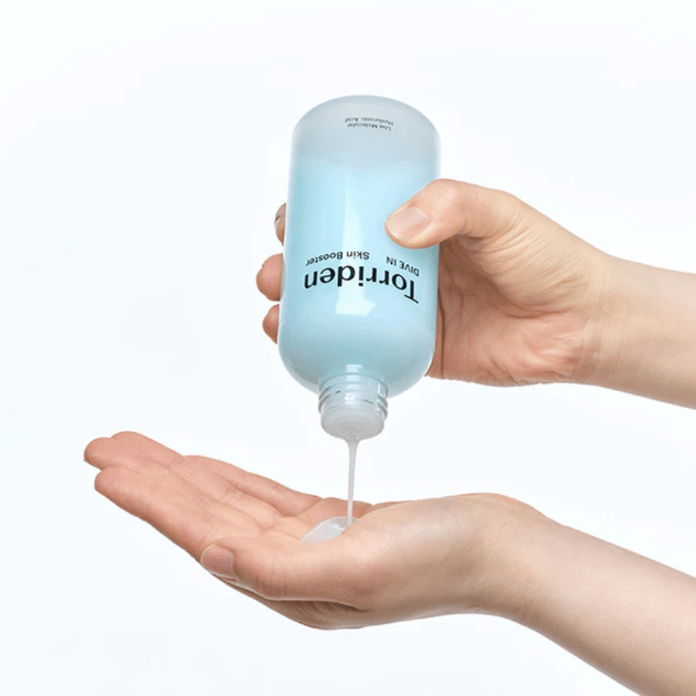 Hænder, der holder en flaske med hudplejeprodukt og doserer en klar væske i håndfladen. Flasken er etiketmærket med "Torriden - DIVE-IN Low Molecule Hyaluronic Acid Skin Booster".