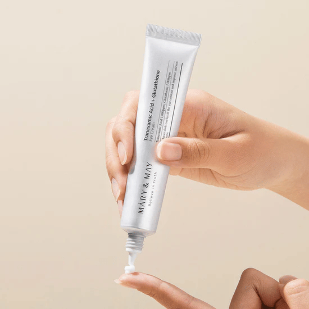 En hånd holder en tube MARY & MAY "Tranexamic Acid + Glutathione Eye Cream". Tuben er hvid, og designet er rent og enkelt, hvilket fremhæver produktets beroligende og lysnende egenskaber for øjenområdet. Produktet påføres nænsomt med fokus på præcision og effektivitet.