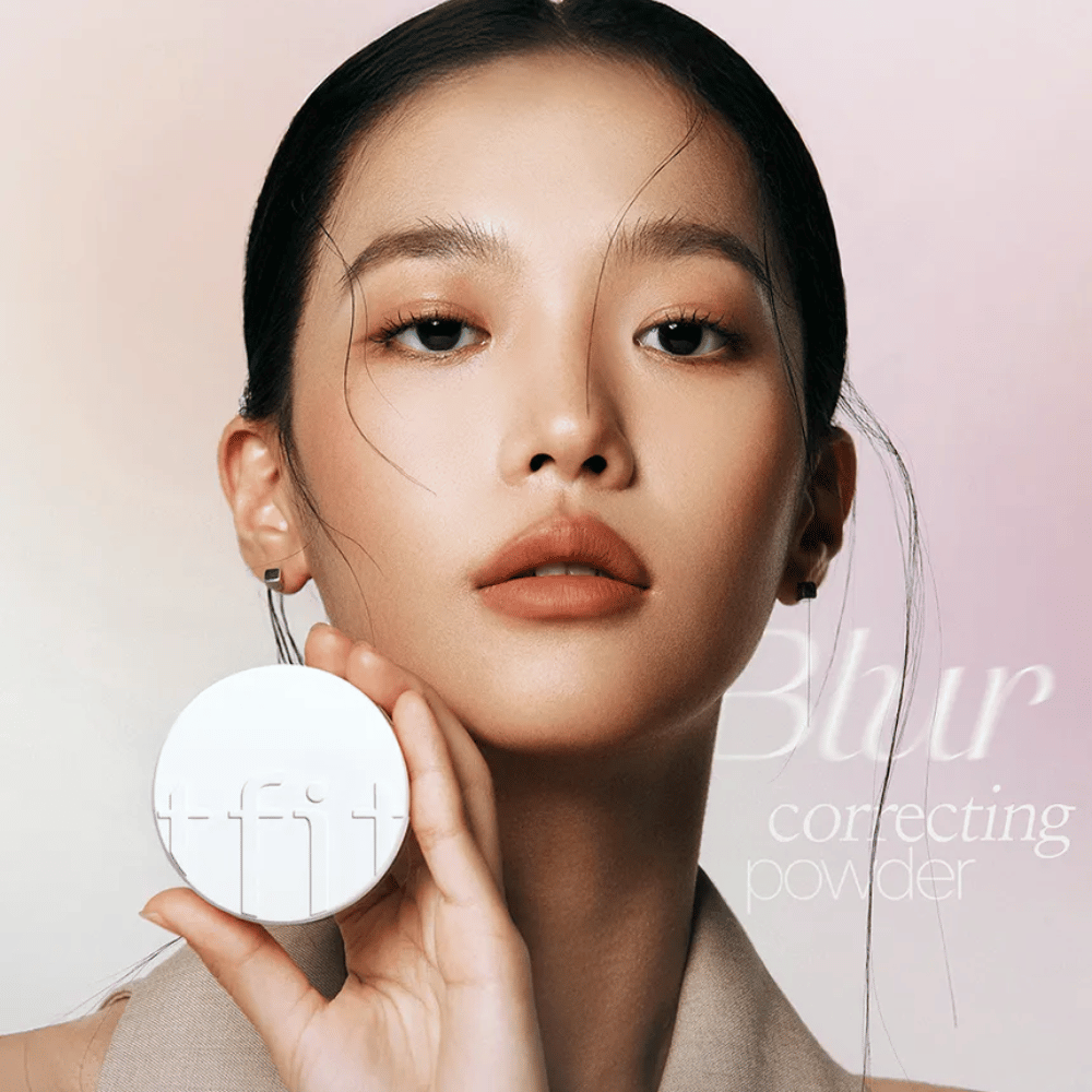 Kvinde med fejlfri makeup holder en rund hvid dåse med TFIT-logo op, med teksten "Blur Correcting Powder" i baggrunden.