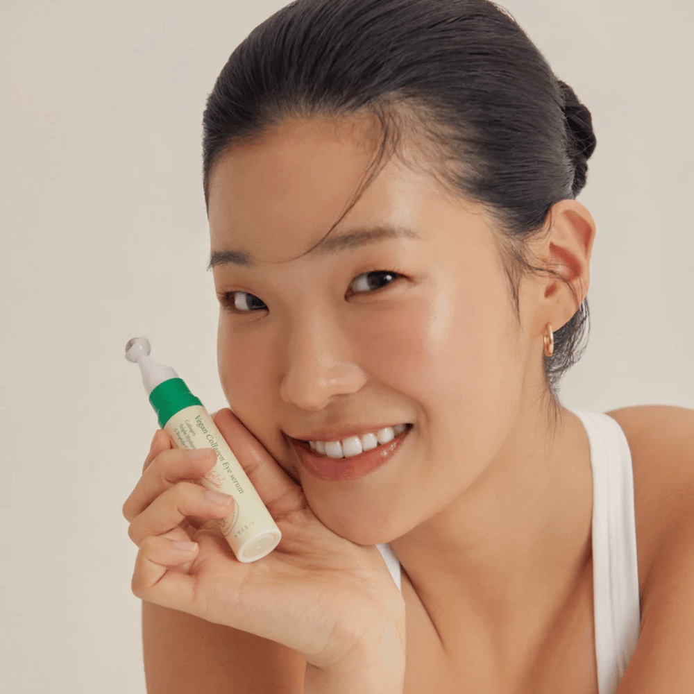 En kvinde med opsat hår og gyldne øreringe holder en AXIS-Y Vegan Collagen Eye Serum 10ml tæt på ansigtet, mens hun smiler blidt. Serummet er designet til at fugte og reducere rynker omkring øjnene, specielt udviklet til følsom hud.