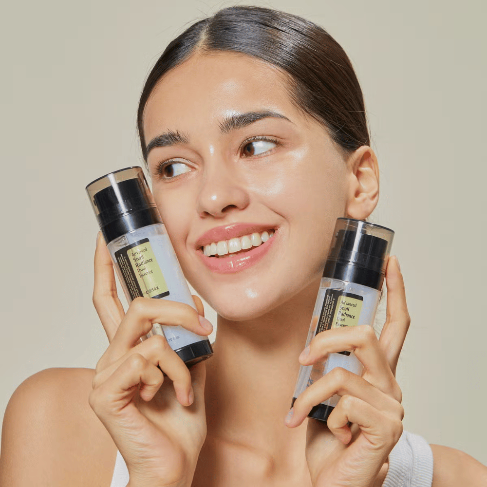 Smilende kvinde med glansfuld hud holder to flasker COSRX Advanced Snail Radiance Dual Essence 80 ml tæt på ansigtet mod en lys baggrund.