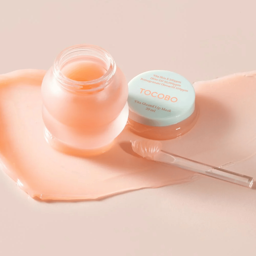 En krukke med "Vita Glazed Lip Mask" fra Tocobo vises. Krukken er gennemsigtig med et lyseturkis låg og indeholder en glansfuld, rosa læbemaske. En lille spatel ligger ved siden af krukken, og en del af masken er spredt ud på underlaget. Produktet er designet til at fugte og pleje læberne.