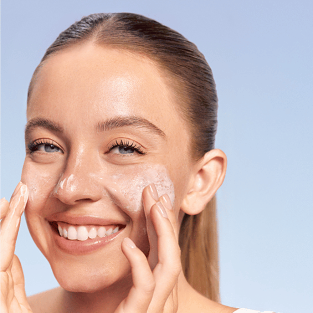 Smilende kvinde påfører LANEIGE Water Bank Gentle Gel Cleanser i skummende form på kinderne, mod blå baggrund.
