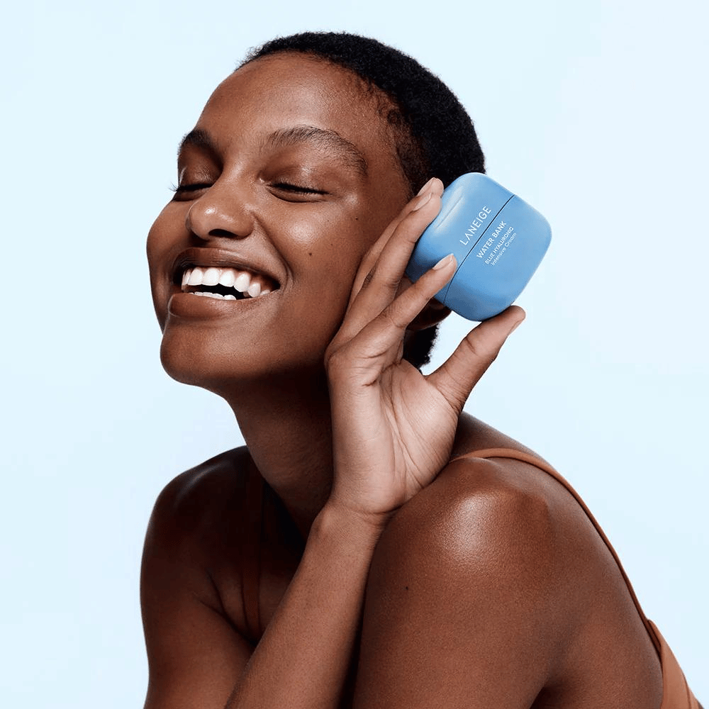 Smilende kvinde holder en blå dåse LANEIGE Water Bank Blue Hyaluronic Cream mod kinden, med lyseblå baggrund.