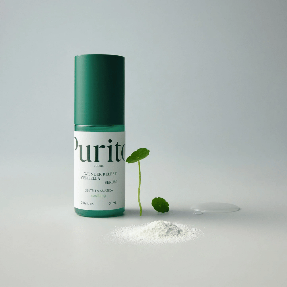 Grøn flaske af Purito Wonder Releaf Centella Serum 60 ml står på et hvidt underlag, omgivet af centellablade, en gennemsigtig serumdråbe og et lille hvidt pulver.