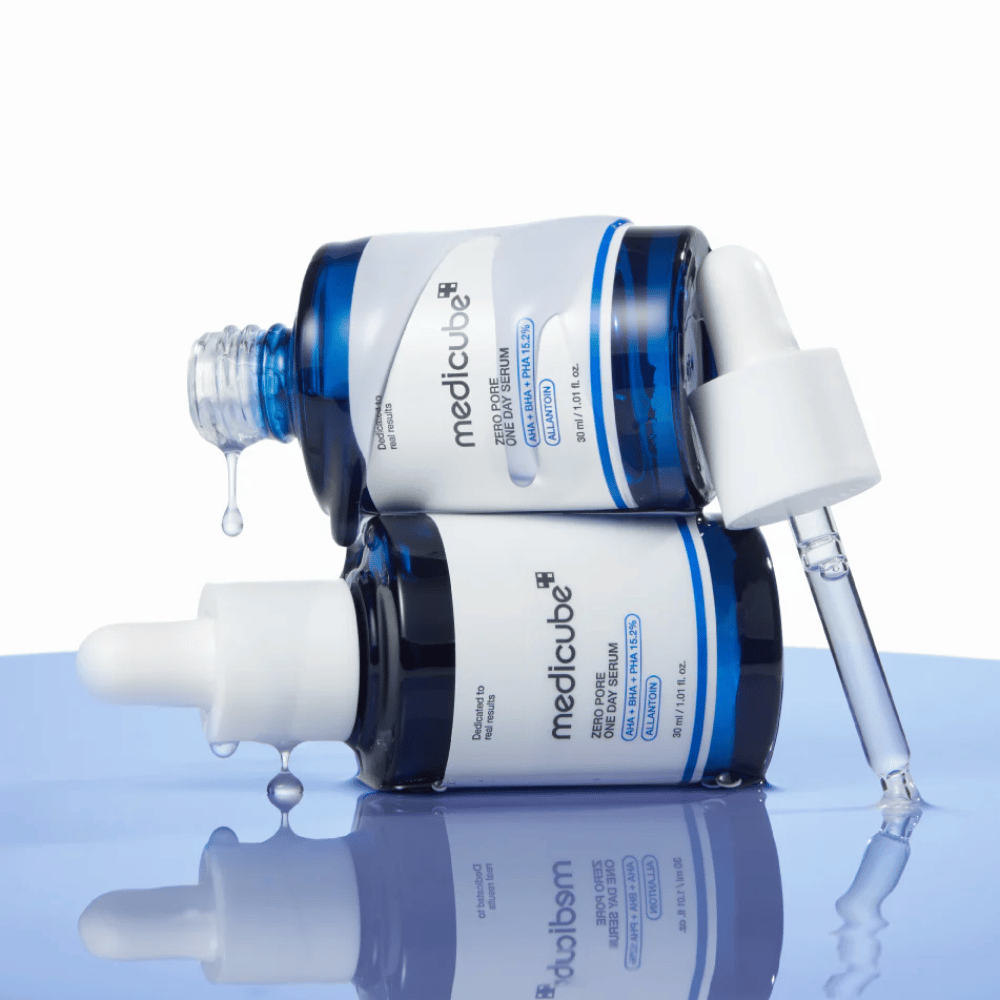 To blå flasker af Medicube Zero Pore One Day Serum liggende mod hinanden, med pipetter og dråber af serum på en reflekterende blå overflade.