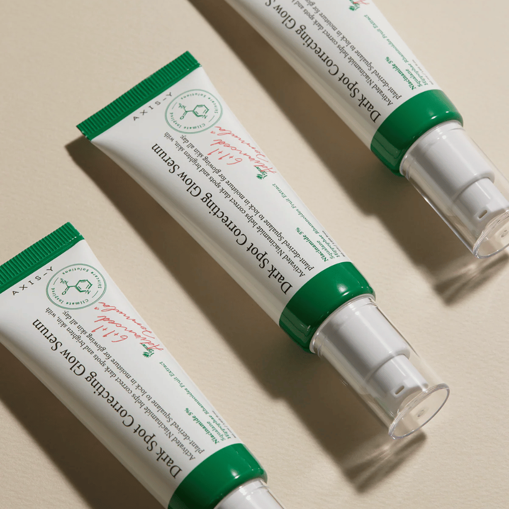 Tre tuber af Axis-Y Dark Spot Correcting Glow Serum med grøn hætte og hvid emballage ligger på en lys overflade.