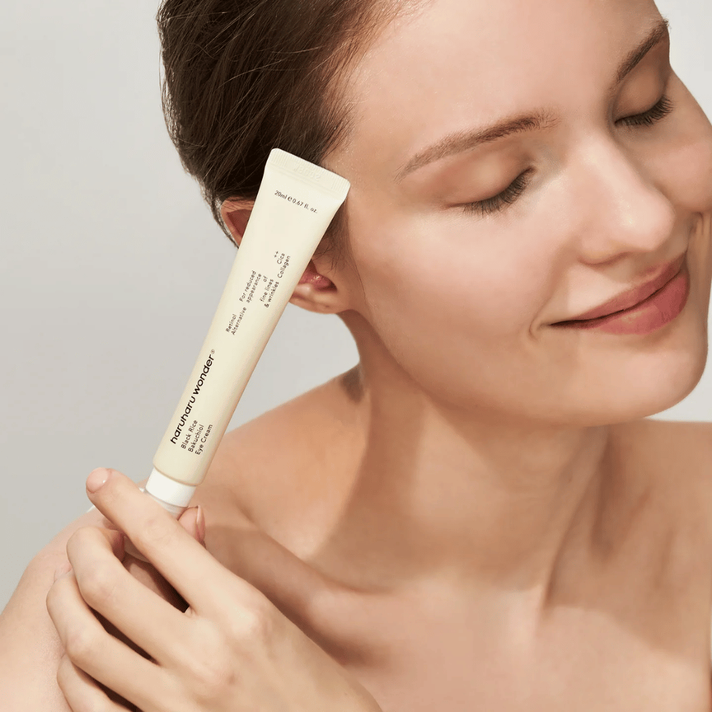 En smilende person med lukkede øjne holder en tube Haruharu Wonder Black Rice Bakuchiol Eye Cream mod tindingen.