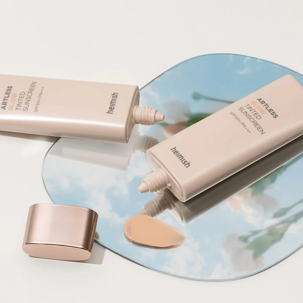En flaske heimish Artless Glow Tinted Sunscreen SPF50+ PA+++ 40ml og en flaske concealer på et spejl, som viser forskellige hudtoner.