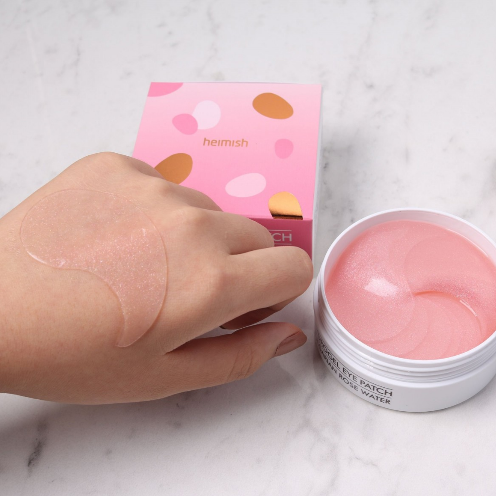 En hånd som holder en lyserød Bulgarian Rose Hydrogel Eye Patch 60stk fra heimish og anvender øjenmaske.