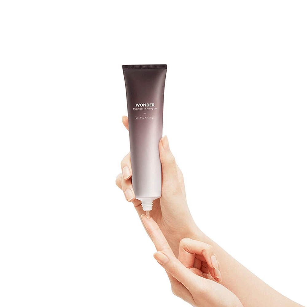 En kvindes hånd, der holder et rør Haruhuru Wonders Black Rice Soft Peeling Gel 100 ml på en hvid baggrund.