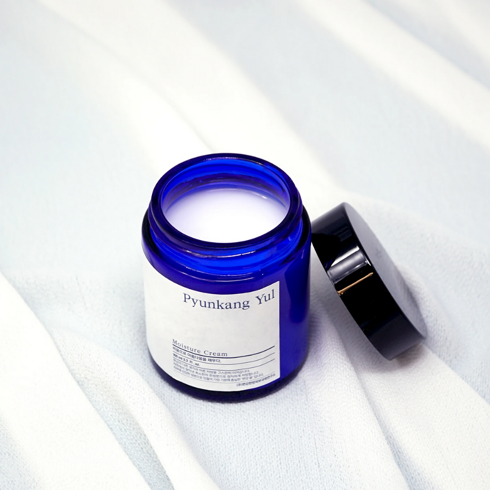 En blå dåse med låg, der sidder ovenpå en hvid klud, der viser Pyunkang Yul's Moisture Cream 100ml fugtgivende egenskaber.
