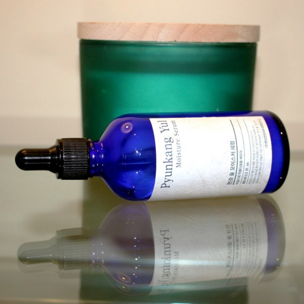 En flaske Pyunkang Yul Moisture Serum 100ml balancerer fugtniveauerne på et bord ved siden af et lys.